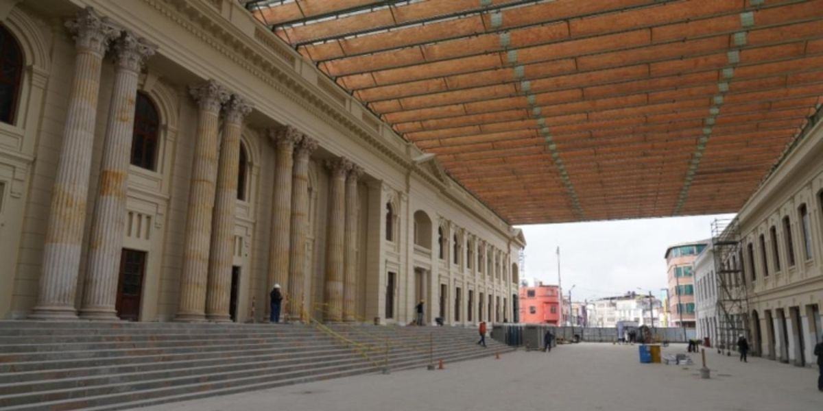 Edificio con columnas y escalinatas bajo una cubierta. Varias personas caminan o permanecen de pie en un patio central.