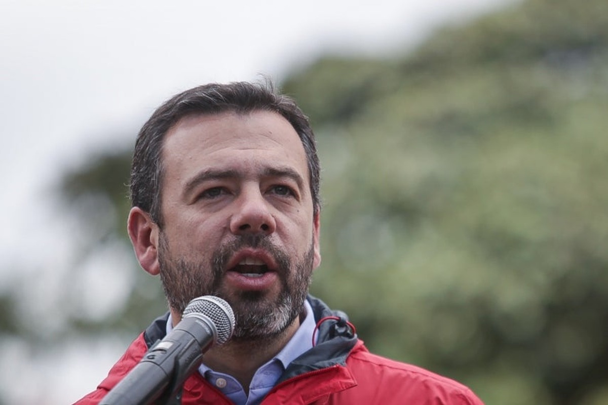 Hombre adulto de barba corta, en chaqueta roja, habla frente a un micrófono plateado al aire libre; fondo desenfocado de vegetación.
