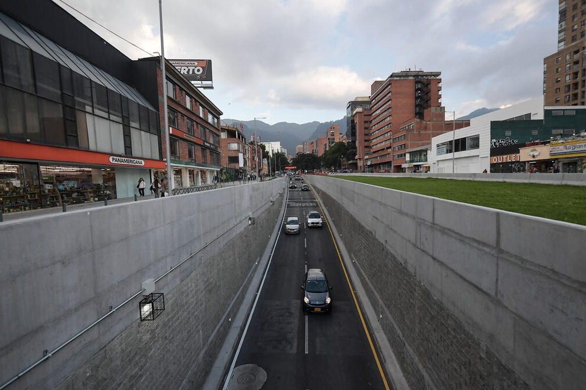 Avenida hundida de dos carriles con varios autos; muros grises a los lados. Arriba hay aceras con peatones, edificios comerciales y de oficinas; se lee “PANAMERICANA” en un letrero.