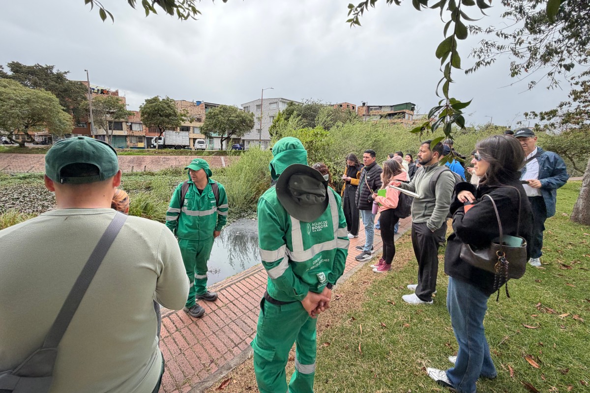 Grupo de personas junto a un canal; varias llevan uniformes verdes. Al fondo, árboles, edificios y cielo nublado.
